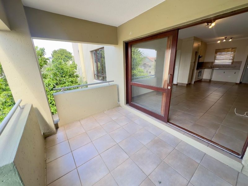 A covered balcony with tiled flooring and a metal balustrade, providing seamless access to the interior living area through a large sliding glass door.