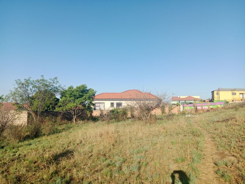 A view of a residential plot featuring natural grass terrain with a boundary wall and neighboring houses in the background.