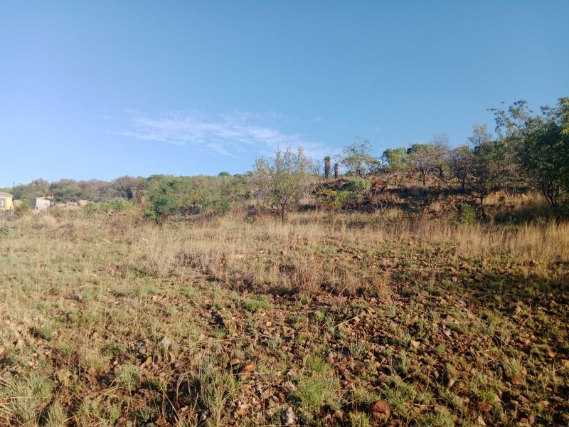A view of a sloped, undeveloped residential plot featuring natural vegetation and rocky terrain under a clear blue sky.