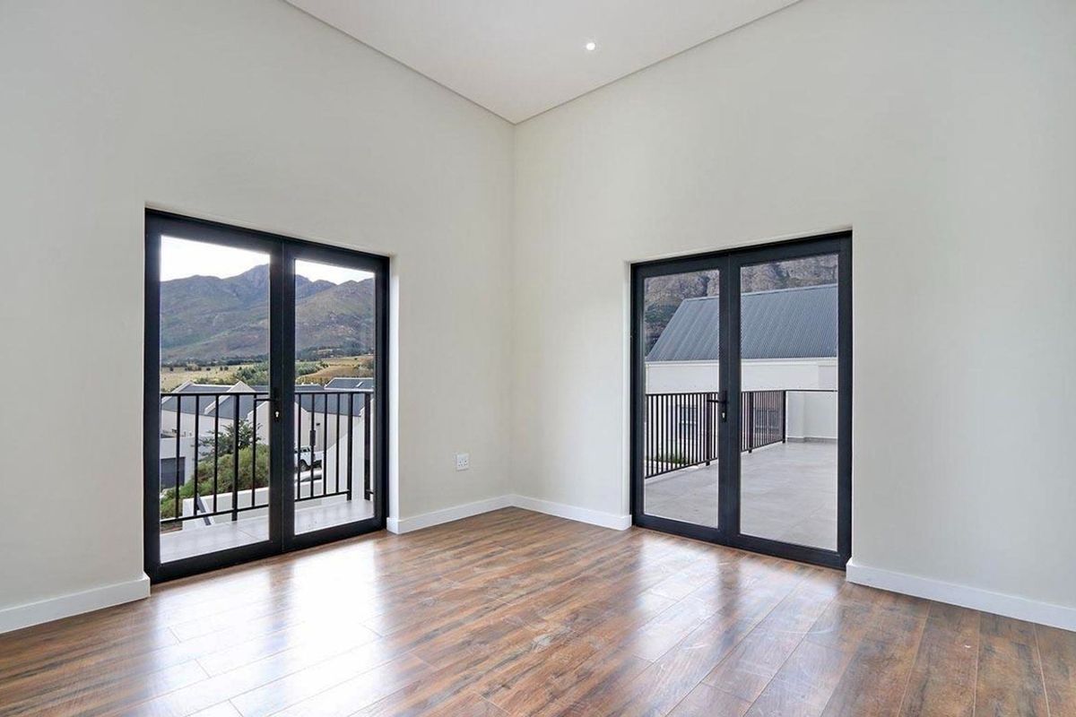 A bright, empty bedroom featuring modern laminate flooring, high ceilings, and dual access points to a balcony with scenic mountain views.
