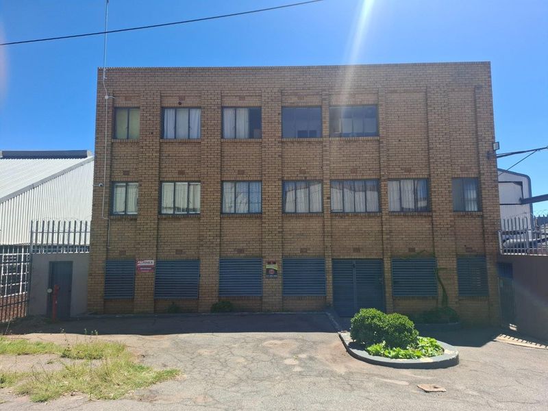 Front elevation of a multi-story industrial brick building featuring security shutters, paved driveway access, and perimeter security fencing.
