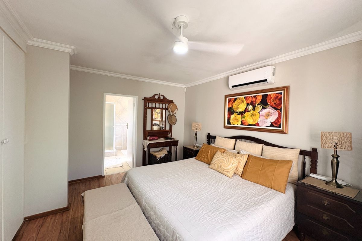A well-maintained bedroom featuring laminate flooring, crown molding, a ceiling fan, and a wall-mounted split-system air conditioner, with an adjoining en-suite bathroom.