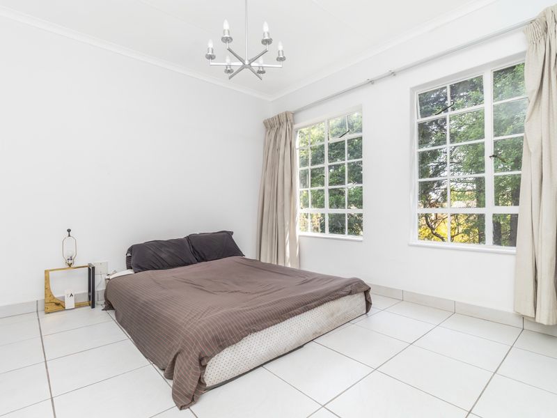A bright bedroom featuring large multi-pane windows, tiled flooring, and a central ceiling light fixture, offering a clean and neutral space.