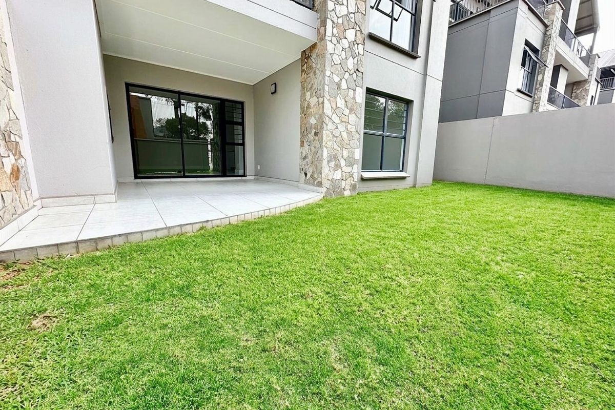 A well-maintained rear garden featuring a covered tiled patio, stone-clad architectural detailing, and modern aluminum-framed sliding doors leading into the apartment.