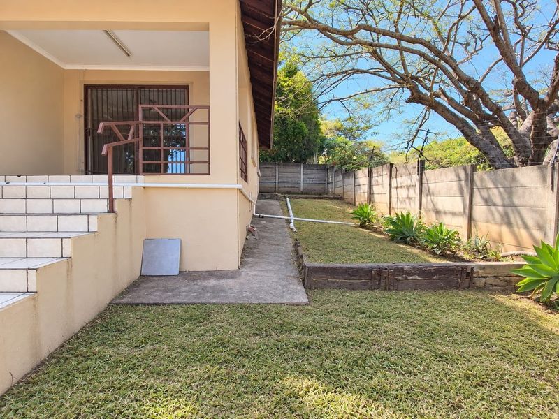 Rear exterior view featuring a tiled patio entrance, concrete pathway, landscaped garden area, and secure precast boundary walling with electric fencing.