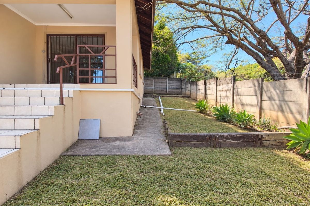 Rear exterior view featuring a tiled patio entrance, concrete pathway, landscaped garden area, and secure precast boundary walling with electric fencing.