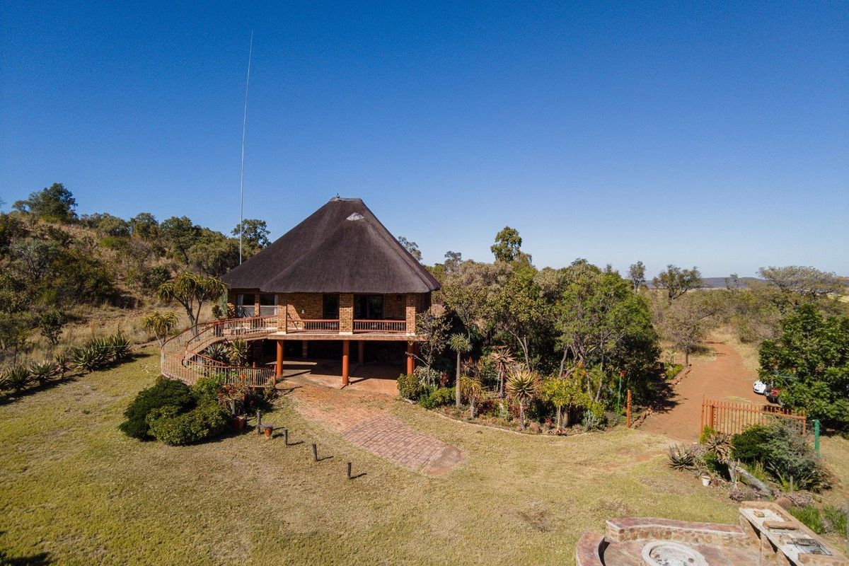 Aerial view of a farm property featuring a raised house with a thatched roof, wrap-around deck, and surrounding natural landscape.