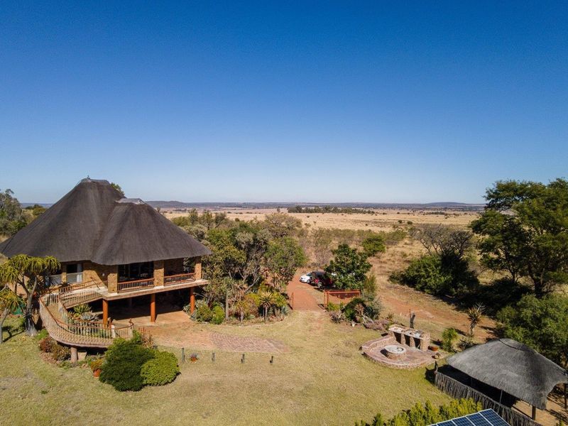 Aerial view of a farm property featuring a thatched-roof house with a raised deck, outdoor braai area, and expansive surrounding landscape.