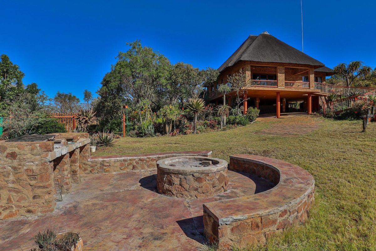 Rear garden area featuring a stone-built fire pit and braai area, overlooking a two-story farmhouse with a thatched roof and elevated deck.