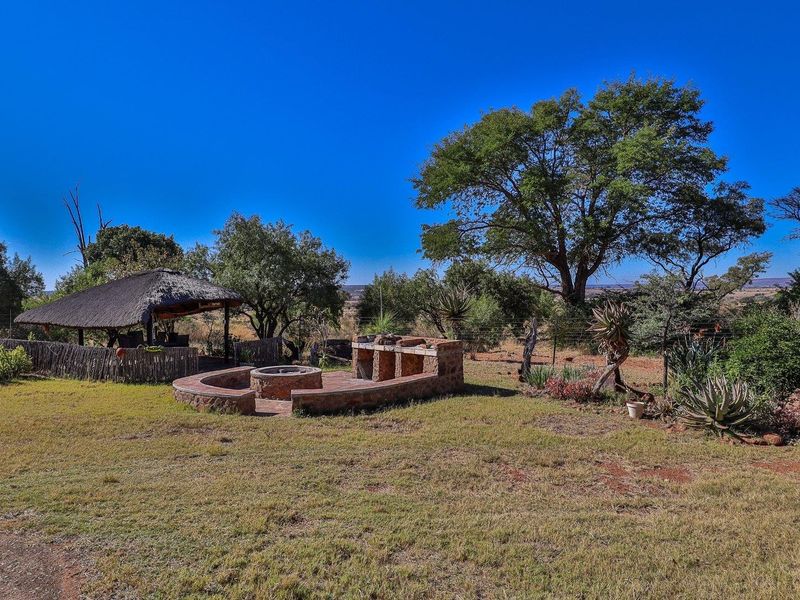 Outdoor entertainment area featuring a thatched lapa, built-in stone braai, and a circular fire pit set within a spacious garden landscape.