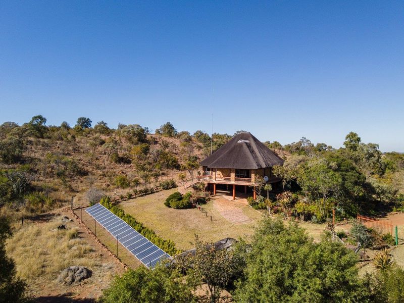 Aerial view of a farm property featuring a thatched-roof dwelling, an elevated deck, and a ground-mounted solar panel array set in a natural landscape.