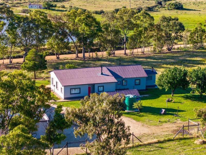 Aerial view of a rural farm property featuring a main dwelling with a metal roof, surrounding green fields, mature trees, and a water storage tank.