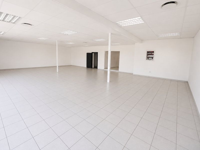 A spacious, empty open-plan commercial office or retail area featuring neutral ceramic floor tiles, a suspended ceiling with recessed lighting, and support columns.
