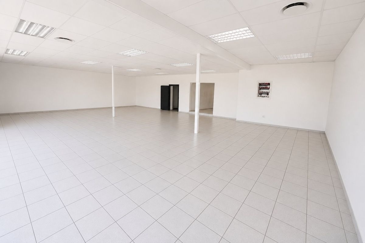 A spacious, empty open-plan commercial office or retail area featuring neutral ceramic floor tiles, a suspended ceiling with recessed lighting, and support columns.