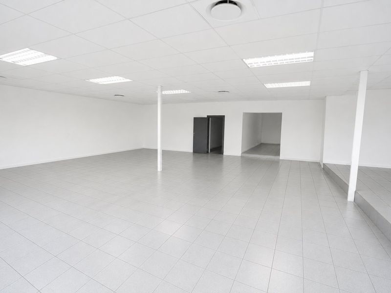 A spacious, vacant open-plan commercial office or retail space featuring neutral ceramic tiling, a suspended ceiling with recessed lighting, and a raised platform section.