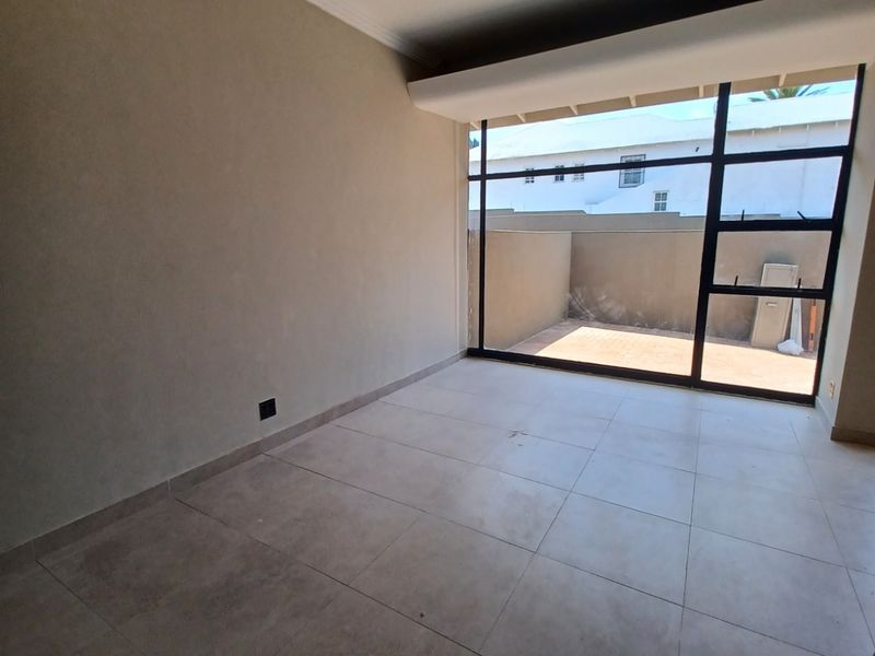 A bright, empty living room featuring large format floor tiles, recessed lighting, and a large black-framed glass door leading to an outdoor patio area.