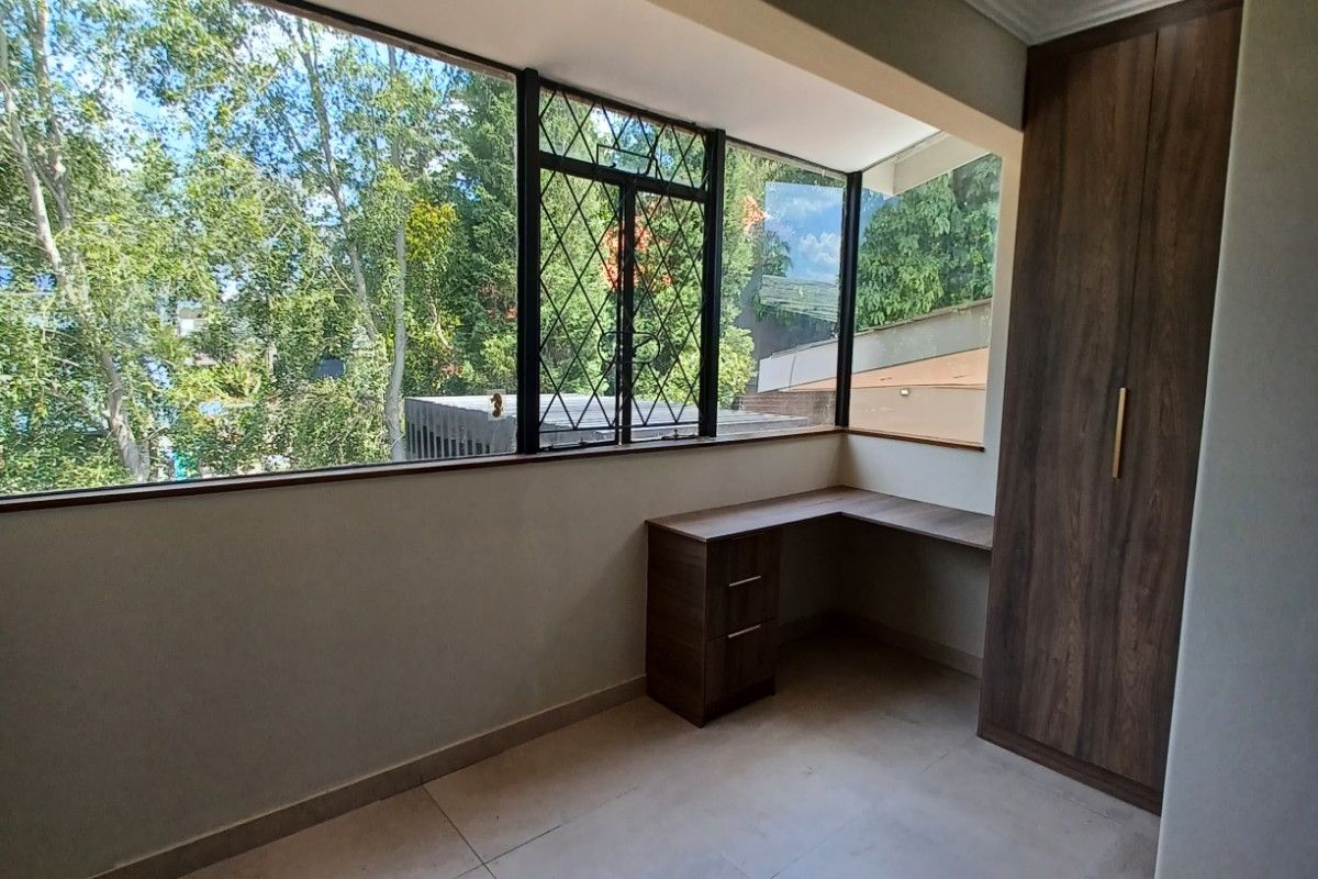 A bright, empty bedroom featuring tiled flooring, a built-in wooden wardrobe, and a custom built-in desk area with large windows overlooking greenery.