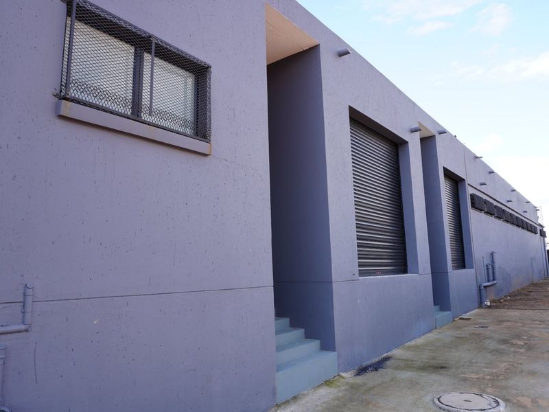 Rear exterior view of a commercial building featuring concrete walls, security roller shutters, and protective window grilles with a concrete apron.