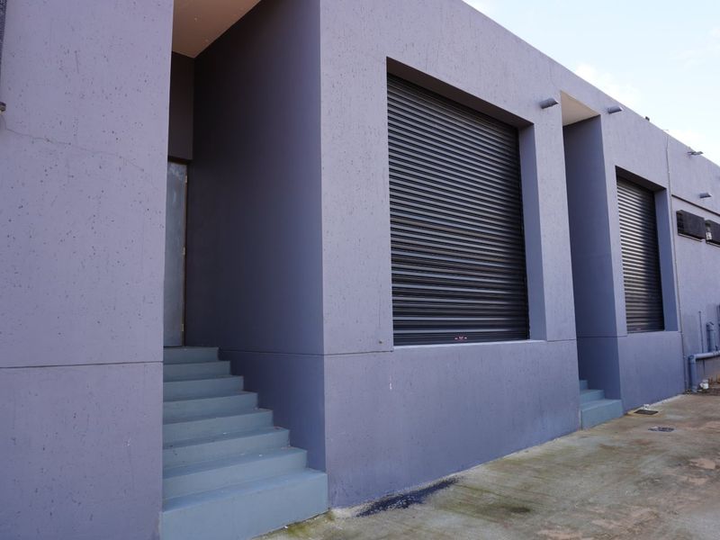 Rear exterior view of a commercial building featuring secure roller shutter doors, concrete access stairs, and external wall-mounted lighting.