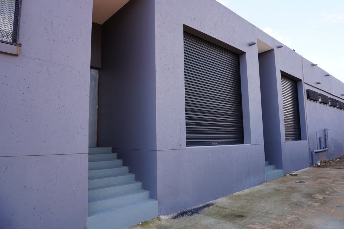 Rear exterior view of a commercial building featuring secure roller shutter doors, concrete access stairs, and external wall-mounted lighting.