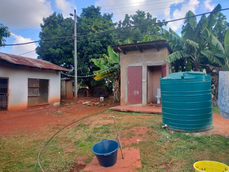 Rear exterior view showing a small outbuilding, water storage tank, and basic utility area with exposed ground.