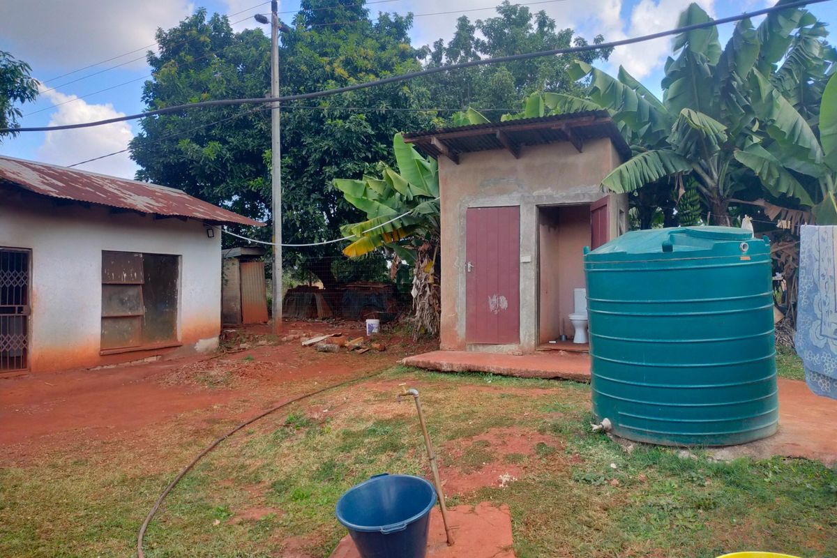 Rear exterior view showing a small outbuilding, water storage tank, and basic utility area with exposed ground.