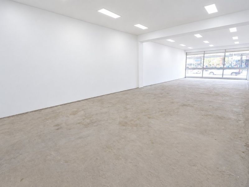 A spacious, empty commercial open-plan unit featuring white walls, recessed ceiling lighting, and a large glass storefront window with unfinished concrete flooring.