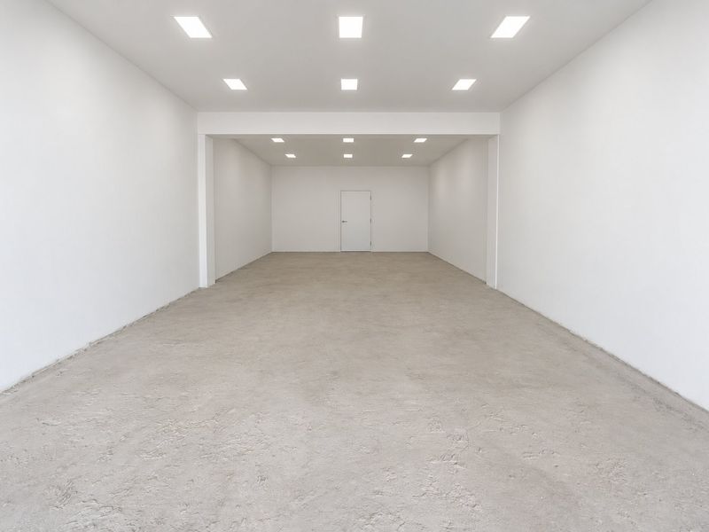 A spacious, empty commercial open-plan area featuring white walls, recessed ceiling lighting, and an unfinished concrete floor.