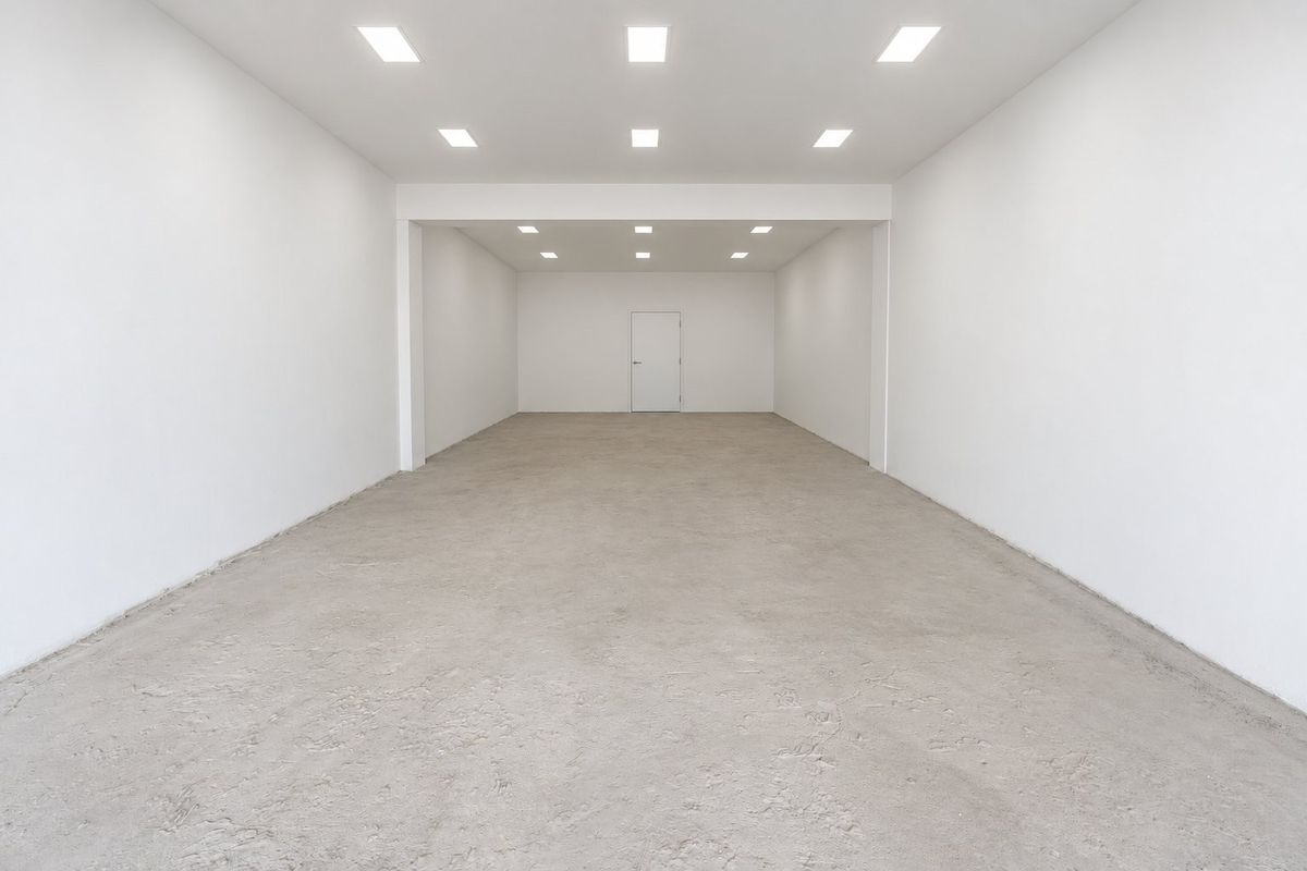 A spacious, empty commercial open-plan area featuring white walls, recessed ceiling lighting, and an unfinished concrete floor.
