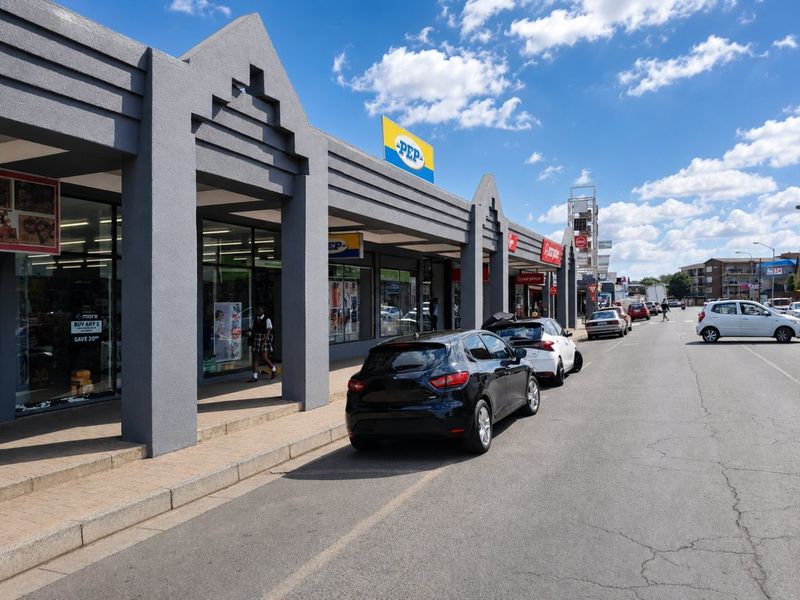 Street-level view of a commercial retail strip center featuring a covered walkway, large display windows, and paved sidewalk access.