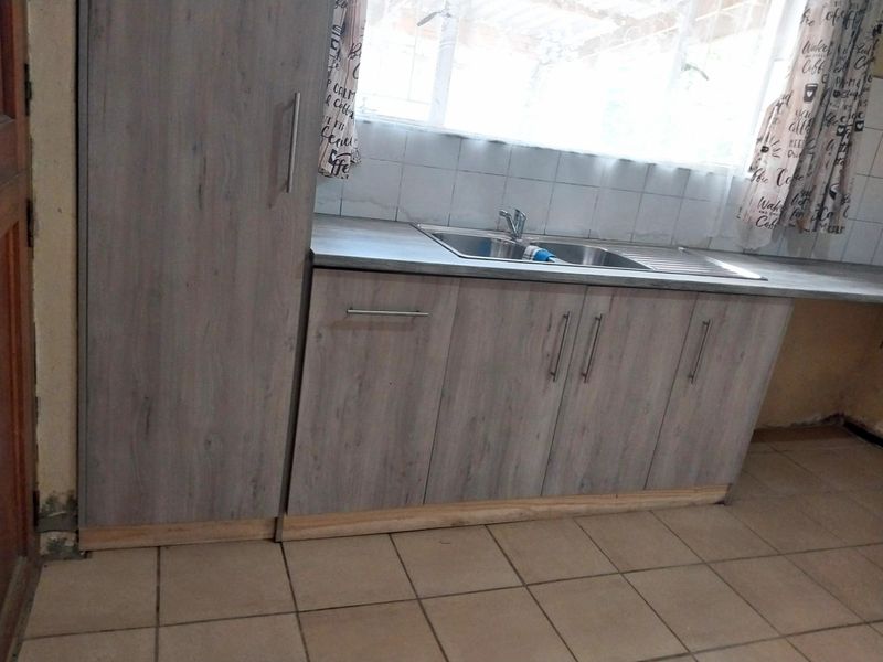 A functional kitchen area featuring light wood-look cabinetry, a stainless steel sink, and tiled flooring, currently requiring minor repairs to baseboards and wall finishes.