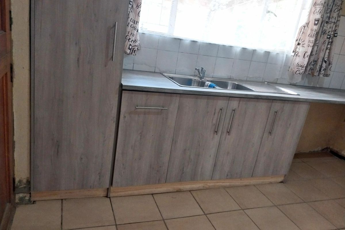 A functional kitchen area featuring light wood-look cabinetry, a stainless steel sink, and tiled flooring, currently requiring minor repairs to baseboards and wall finishes.