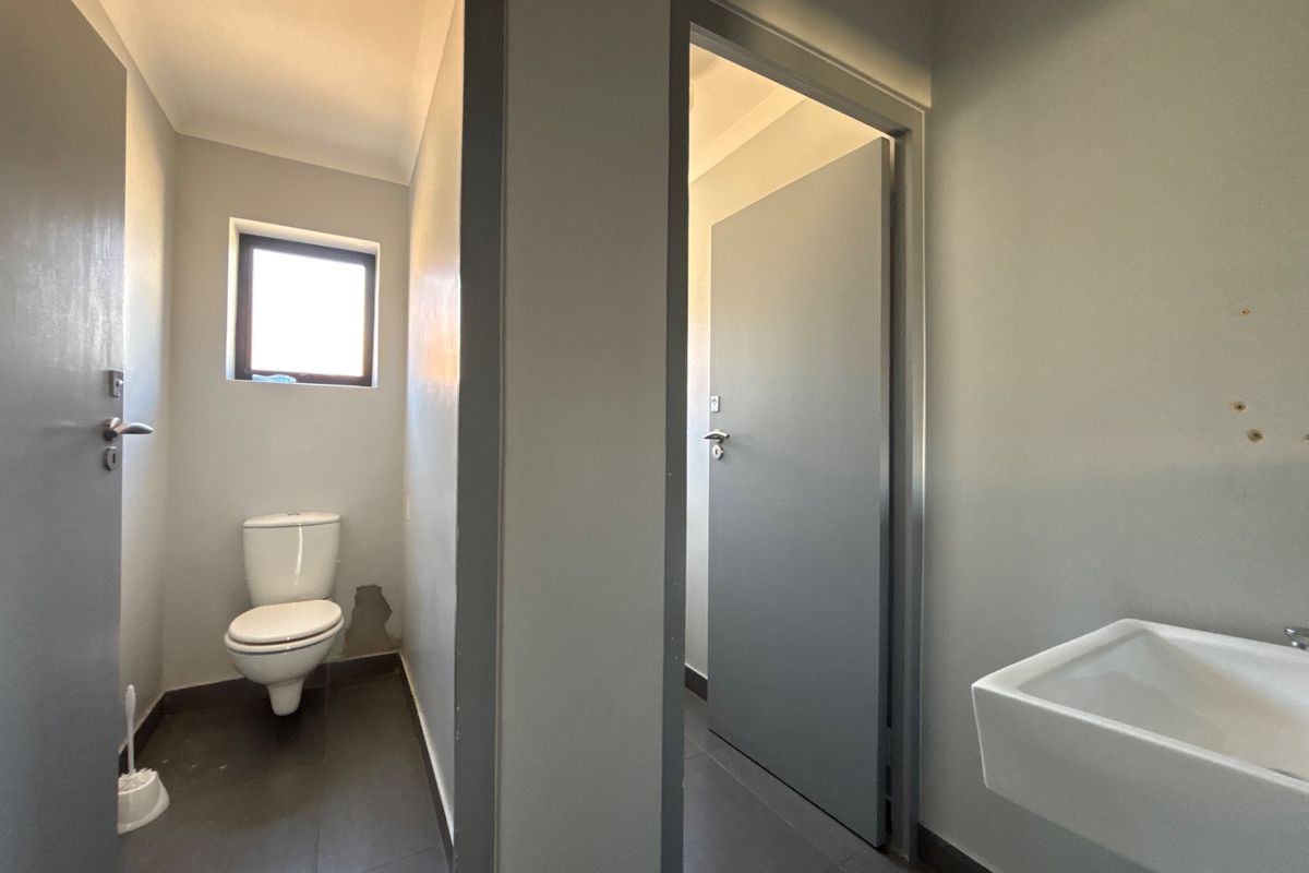 A functional office bathroom featuring a toilet, wall-mounted basin, and tiled flooring, currently showing signs of wear on the wall finishes.