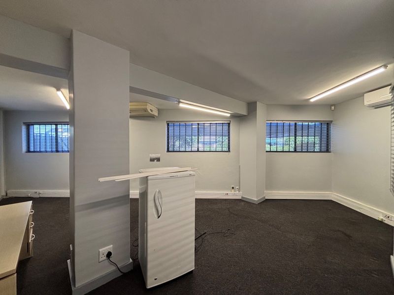 A spacious, open-plan office area featuring neutral walls, carpeted flooring, multiple windows with venetian blinds, and integrated air conditioning units.