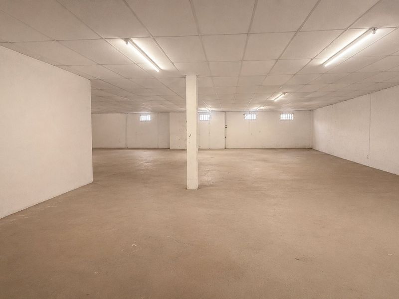 A large, open-plan commercial utility or storage space featuring concrete flooring, a suspended ceiling with fluorescent lighting, and small high-level windows.
