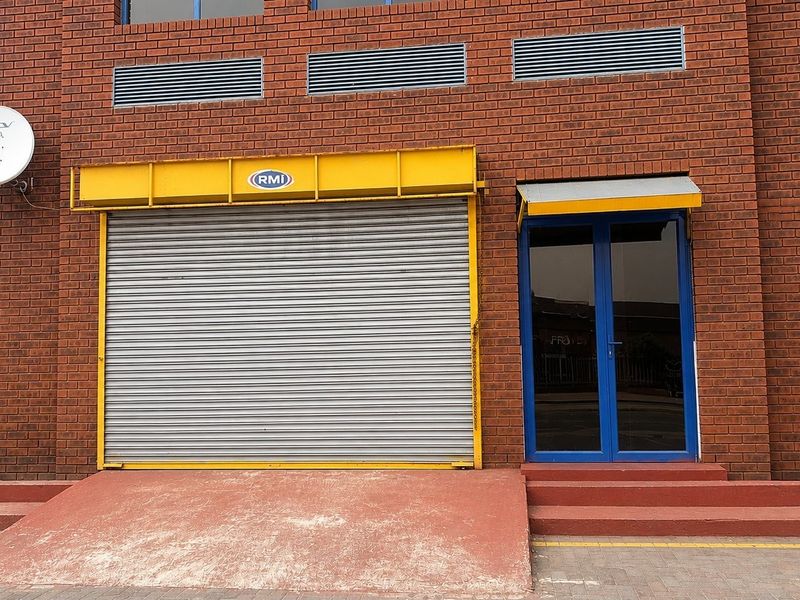 Commercial building exterior featuring a red brick facade, secure roller shutter door, glass entrance door, and a concrete access ramp.