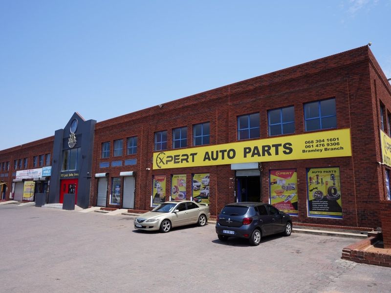 A commercial brick building featuring large display windows, metal roller shutter doors, and a paved parking area, currently occupied by an auto parts store.