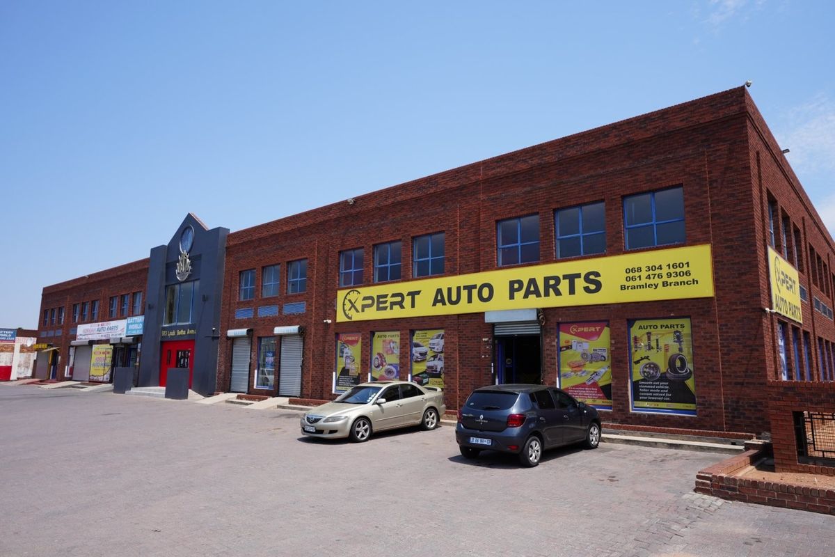 A commercial brick building featuring large display windows, metal roller shutter doors, and a paved parking area, currently occupied by an auto parts store.