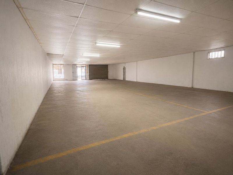 A spacious, empty commercial garage or warehouse area featuring concrete flooring, a suspended ceiling with fluorescent lighting, and a secure roller shutter entrance.