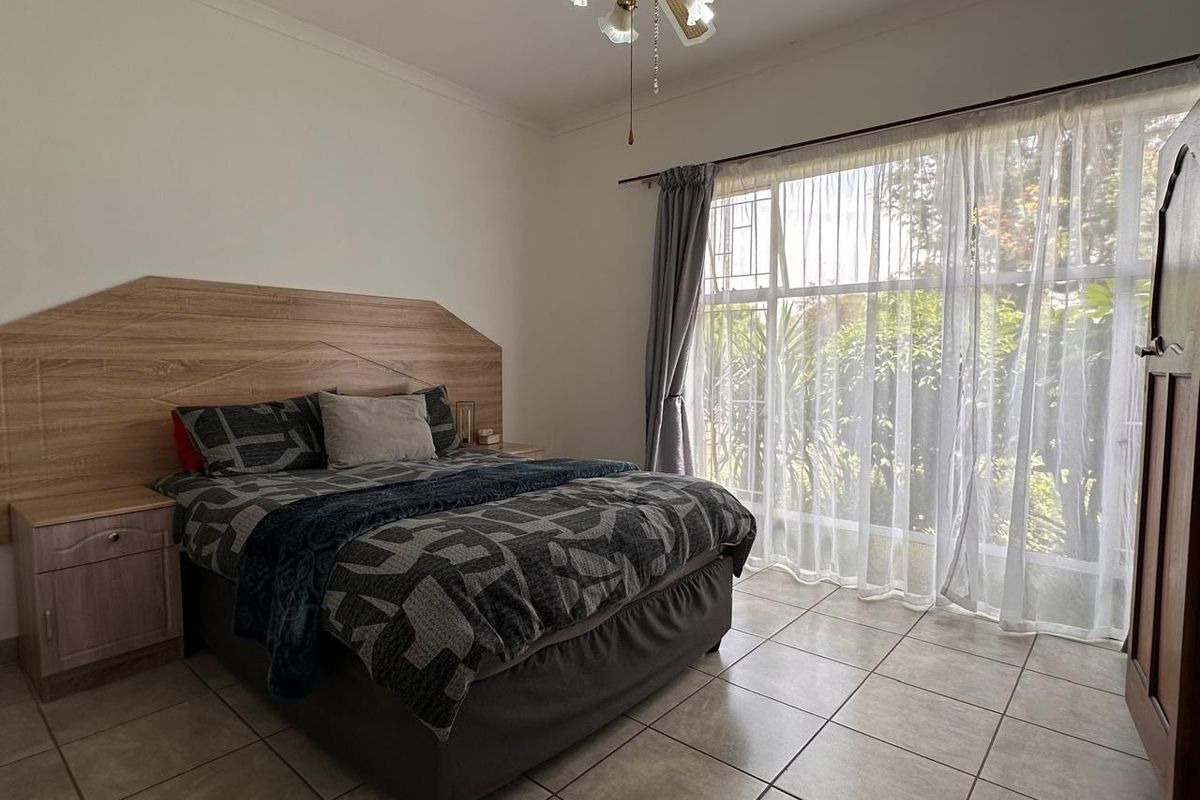 A well-lit bedroom featuring tiled flooring, a built-in headboard with integrated bedside cabinets, and a large window overlooking the garden.