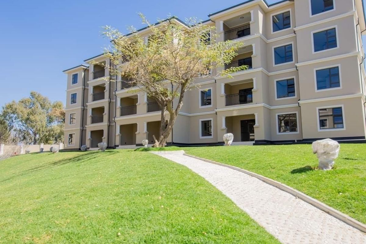 A well-maintained multi-story apartment building exterior featuring a paved walkway, landscaped lawn, and private balconies for each unit.