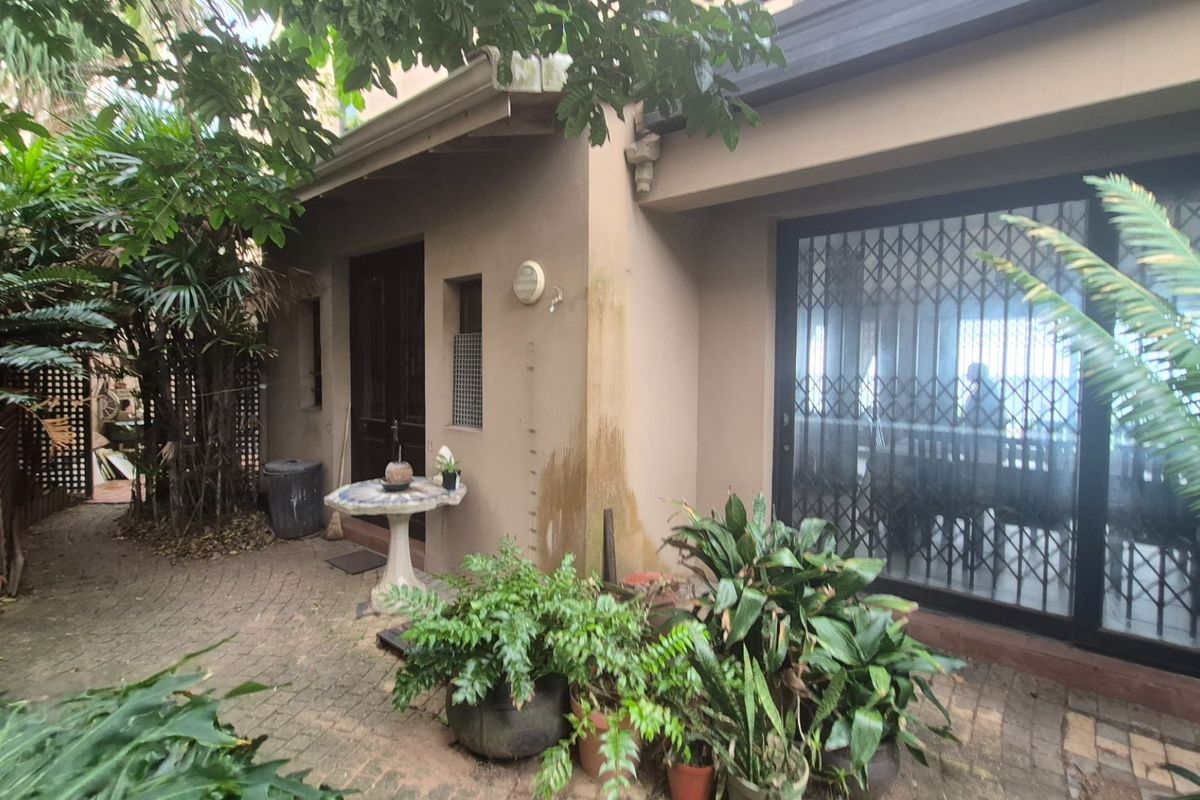 A rear exterior view of the property featuring a paved patio area, sliding glass doors with security gates, and established garden foliage.