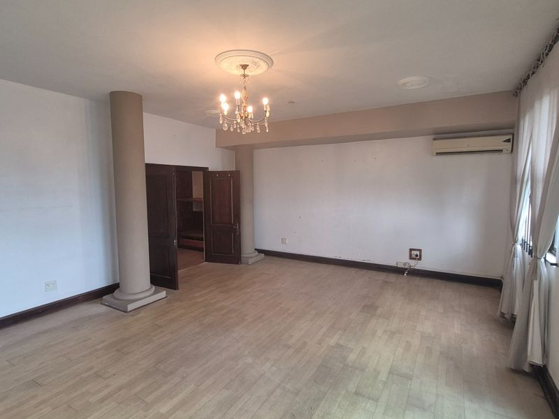 Spacious living area featuring laminate flooring, structural columns, a decorative ceiling rose with chandelier, and a wall-mounted air conditioning unit.