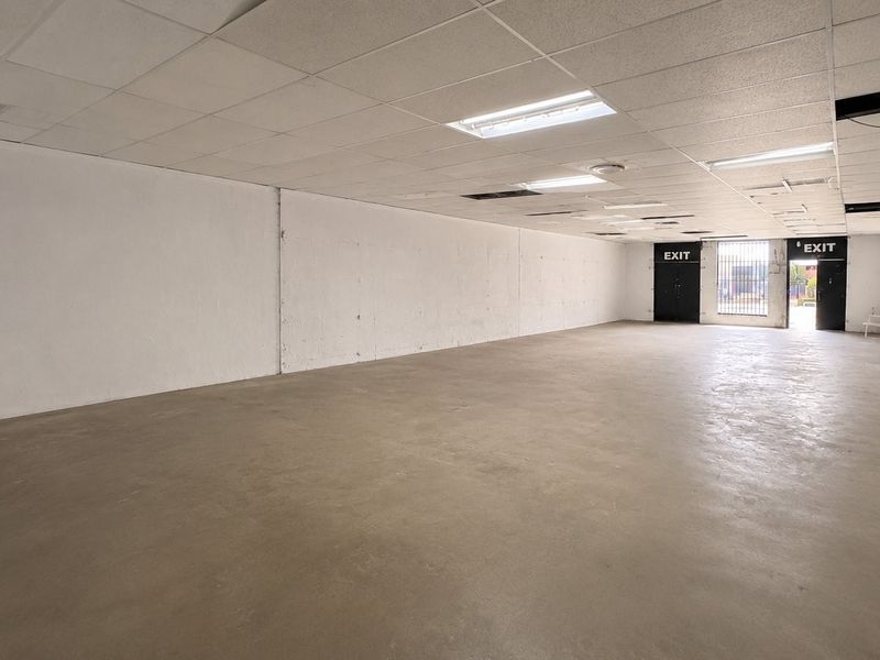 A large, vacant open-plan commercial space featuring concrete flooring, a suspended ceiling with recessed lighting, and multiple exit points.