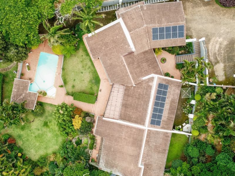 Aerial view of the property featuring a tiled roof with solar panels, a swimming pool, paved patio areas, and established garden landscaping.