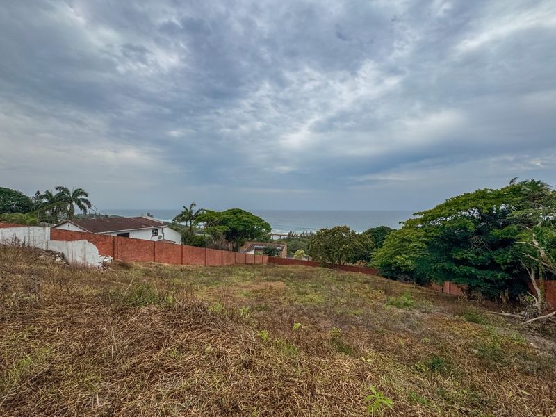 A vacant residential plot featuring a sloping landscape and a scenic ocean view, enclosed by a brick boundary wall.