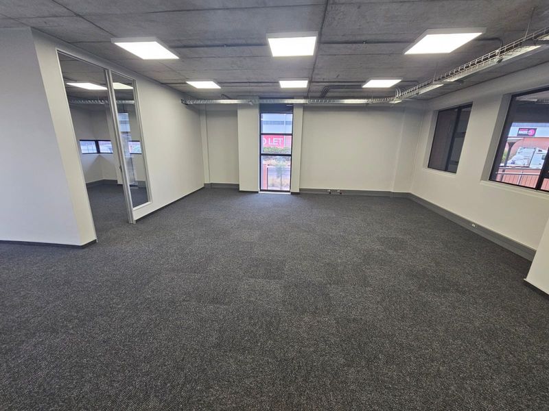 Modern open-plan office space featuring exposed concrete ceilings, recessed LED lighting, carpeted floors, and a glass-partitioned side office.