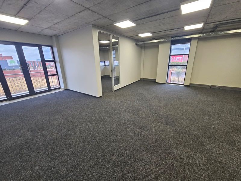Modern open-plan office space featuring exposed concrete ceilings, recessed LED lighting, carpeted floors, and large windows providing ample natural light and balcony access.