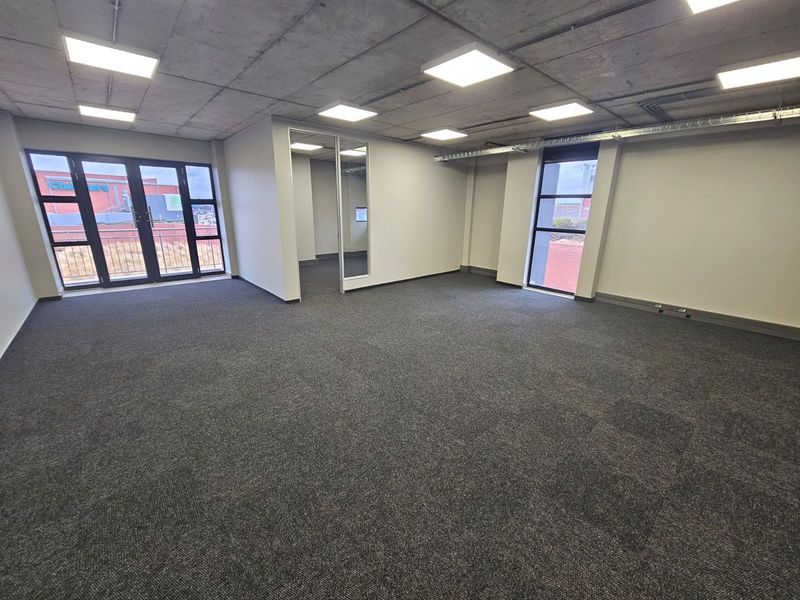 A modern, open-plan office space featuring exposed concrete ceilings, recessed LED lighting, carpeted floors, and large windows providing natural light.