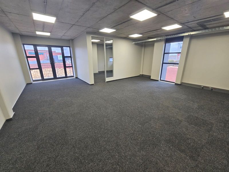 A modern, open-plan office space featuring exposed concrete ceilings, recessed LED lighting, carpeted flooring, and large windows providing natural light.